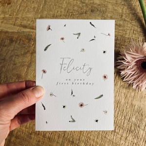 May include: A white card with a floral pattern and the text "Felicity on your first birthday".