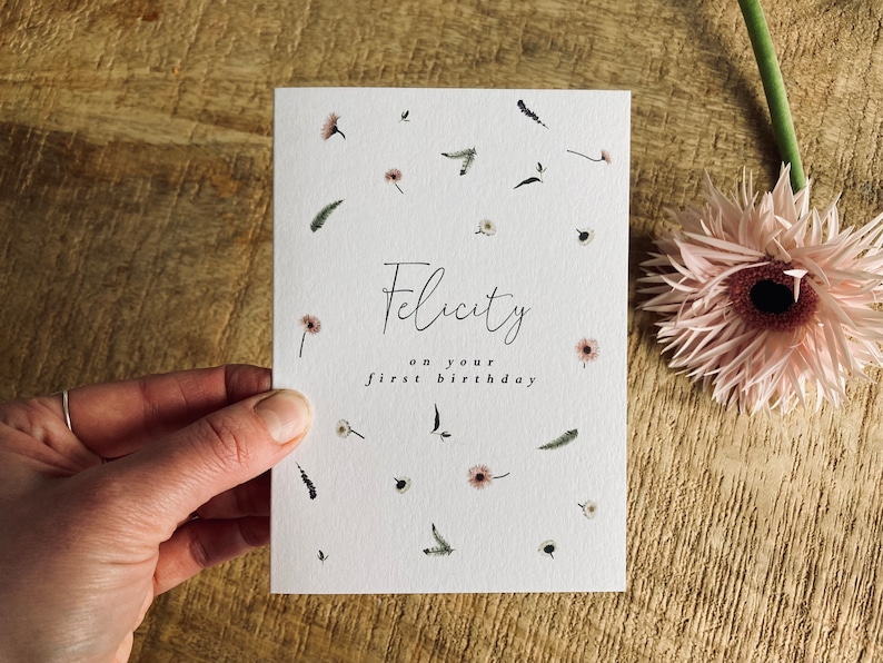May include: A white card with a floral pattern and the text "Felicity on your first birthday".
