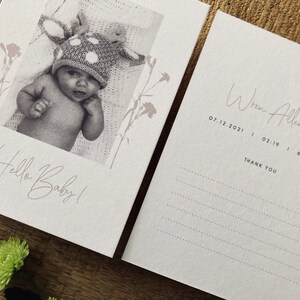 Op de afbeelding: Een witte kaart met een zwart-witfoto van een baby die een stippenhoed draagt. De tekst "Hello Baby!" staat in cursief op de kaart. Een tweede witte kaart heeft de tekst "Wren Allenford 07.12.2021 | 02:19 | 6 LBS 9OZ THANK YOU" in cursief.
