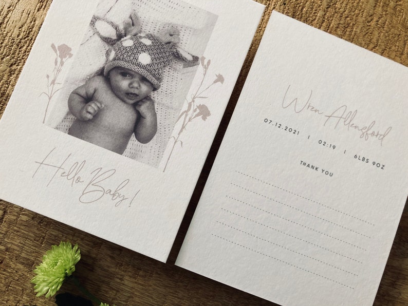 Op de afbeelding: Een witte kaart met een zwart-witfoto van een baby die een stippenhoed draagt. De tekst "Hello Baby!" staat in cursief op de kaart. Een tweede witte kaart heeft de tekst "Wren Allenford 07.12.2021 | 02:19 | 6 LBS 9OZ THANK YOU" in cursief.