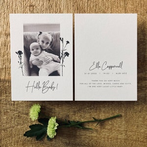 Op de afbeelding: Een zwart-witfoto van twee kinderen, een baby en een peuter, die op een bank zitten. De foto is omlijst met een witte rand en bloemmotieven. De tekst "Hello Baby!" staat onder de foto. De tekst "Ella Coppervell 31.01.2022 | 19:02 | 8LBS 8OZ" staat aan de rechterkant van de kaart. De tekst "Hartelijk dank voor al de liefde, wensen, kaarten en cadeaus. Ik ben een heel gelukkig klein baby." staat onder de datum en het gewicht.