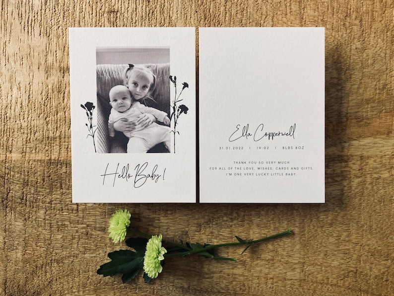 Op de afbeelding: Een zwart-witfoto van twee kinderen, een baby en een peuter, die op een bank zitten. De foto is omlijst met een witte rand en bloemmotieven. De tekst "Hello Baby!" staat onder de foto. De tekst "Ella Coppervell 31.01.2022 | 19:02 | 8LBS 8OZ" staat aan de rechterkant van de kaart. De tekst "Hartelijk dank voor al de liefde, wensen, kaarten en cadeaus. Ik ben een heel gelukkig klein baby." staat onder de datum en het gewicht.