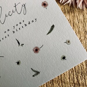 May include: A white card with the text "Felicity on your first birthday" in black script. The card is decorated with small, delicate floral and feather illustrations.
