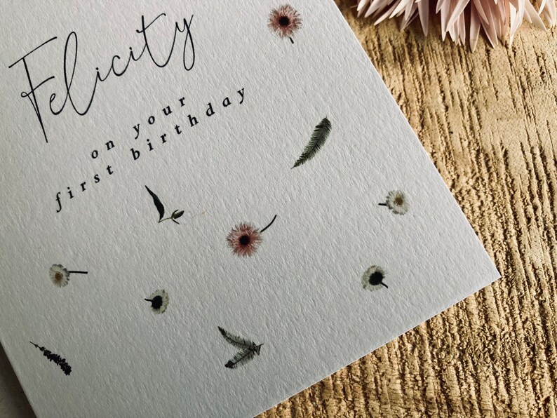 May include: A white card with the text "Felicity on your first birthday" in black script. The card is decorated with small, delicate floral and feather illustrations.