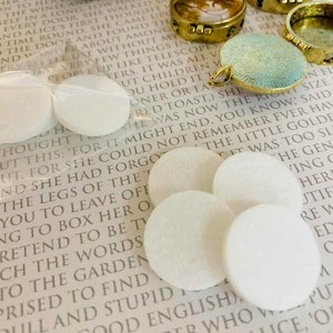 Puede incluir: Seis almohadillas redondas de fieltro blanco, probablemente para usar en manualidades o fabricación de joyas, están dispuestas en una página de un libro con texto. El texto es del libro "Las aventuras de Alicia en el país de las maravillas" de Lewis Carroll.