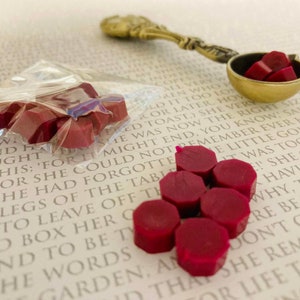 Puede incluir: Primer plano de una bolsa de sellos de cera roja, con algunos sellos esparcidos en una página de un libro. Los sellos son hexagonales y tienen un acabado brillante y lustroso.