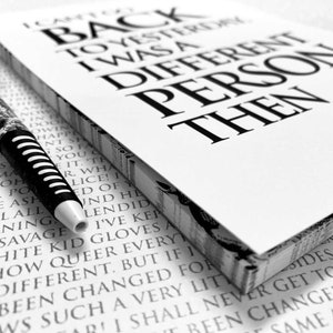 Op de afbeelding: Een zwart-wit notitieboekje met de tekst "Ik kan niet terug naar gisteren, ik was toen een ander persoon". Een zwart-witte pen ligt op het notitieboekje.