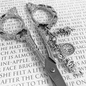 May include: A pair of antique silver scissors with ornate handles and a decorative chain with charms, including a rose, a clock, and a bird, on a page of a book.
