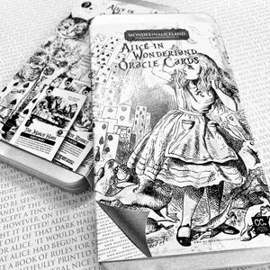 Op de afbeelding: Zwart-wit Alice in Wonderland Oracle Cards in een blikken doos. De doos bevat illustraties van Alice, de Cheshire Cat en andere personages. De kaarten zijn zichtbaar in de doos.