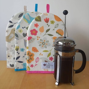 May include: Three colourful tea cosy covers with a bird and floral pattern. The covers are made of fabric and have a pink, blue and white trim. A silver French press coffee maker with dark coffee inside is sitting on a wooden table.