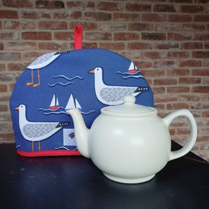 May include: A white ceramic teapot with a blue and white tea cozy featuring a seagull pattern. The tea cozy has a red trim.