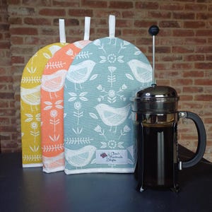 May include: Three fabric coffee pot cosies in yellow, orange, and blue with a white bird and floral pattern. The cozies have a white loop handle. A silver French press with black handle and black lid is in the foreground.