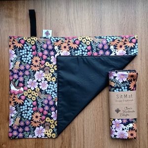 May include: A floral dog sit mat with a black backing, featuring a pattern of colorful flowers and dog illustrations. The mat is shown unfolded and folded, with a black handle and a tag. A smaller, folded mat is also shown.