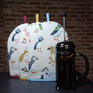 May include: Three white cotton pot holders with a blue trim and a pattern of colourful puffins. The pot holders are in front of a silver French press coffee maker with dark coffee inside.