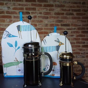 May include: Two French presses with black handles and a tea cozy set. The tea cozies are white with a blue trim and feature a whimsical animal print. One press is silver, the other is gold.