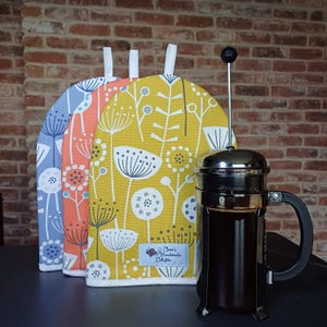 May include: Three yellow, orange and blue fabric pot holders with a white floral pattern. The pot holders have a white trim and a white hanging loop. A silver French press with a black handle is in the foreground. The French press is filled with coffee. A label on the yellow pot holder reads "Clare's Handmade Delights."