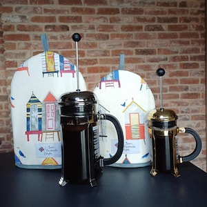 May include: Two French press coffee makers with black handles and glass carafes. The coffee makers are sitting on a black surface. The coffee makers are covered with white fabric covers with a colourful beach hut design. The fabric covers have the text "Clara's Handmade Delights" printed on them.