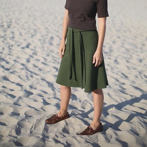 Könnte beinhalten: Eine Frau trägt ein dunkelbraunes Hemd mit Kragen und einen dunkelgrünen Rock mit Gürtel. Sie steht an einem Sandstrand mit dem Meer im Hintergrund. Sie trägt eine Sonnenbrille und blickt zum Himmel.