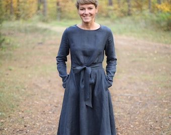 May include: A woman wearing a dark blue linen dress with a tie belt, standing in a forest setting. She is wearing brown ankle boots.