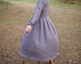 May include: A woman wearing a long, flowing, gray linen dress with a gathered skirt and long sleeves. She is standing in a forest with fall foliage in the background.