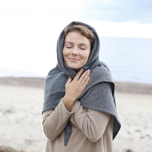 Puede incluir: Una mujer con una bufanda de punto gris y un suéter beige con una falda gris a juego. Está de pie en una playa de arena.