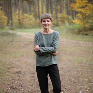 May include: A woman wearing a green long-sleeved shirt and black pants stands in a forest setting. The trees are in the background and the ground is covered in pine needles.