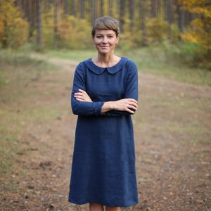 May include: A woman wearing a long-sleeved, navy blue dress with a rounded collar. The dress falls to the mid-calf and is paired with dark brown ankle boots. The background is a blurred forest scene.
