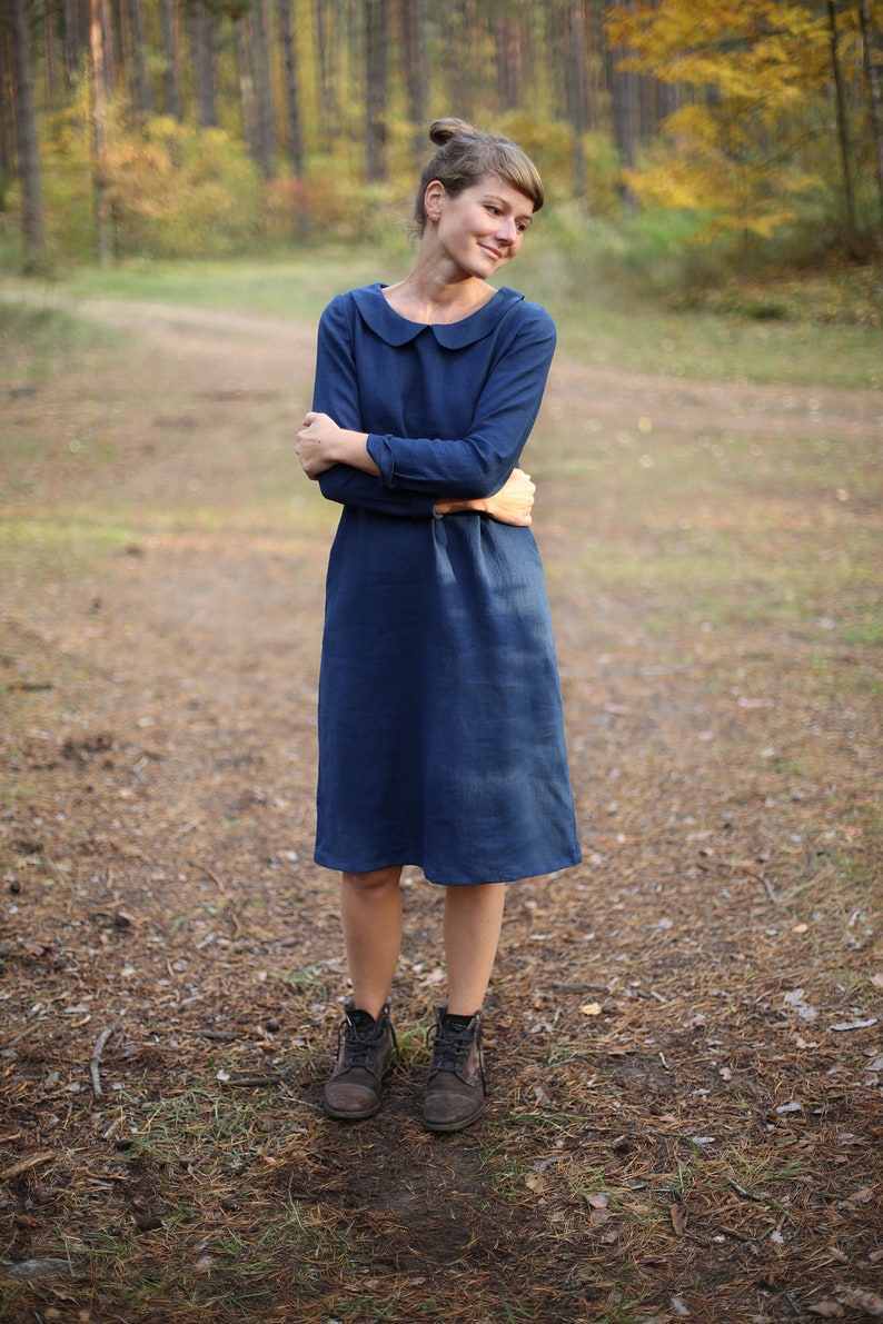 May include: A long-sleeved, navy blue dress with a Peter Pan collar. The dress falls to the mid-calf and is paired with dark brown ankle boots. The background features a forest setting with trees and fallen leaves.
