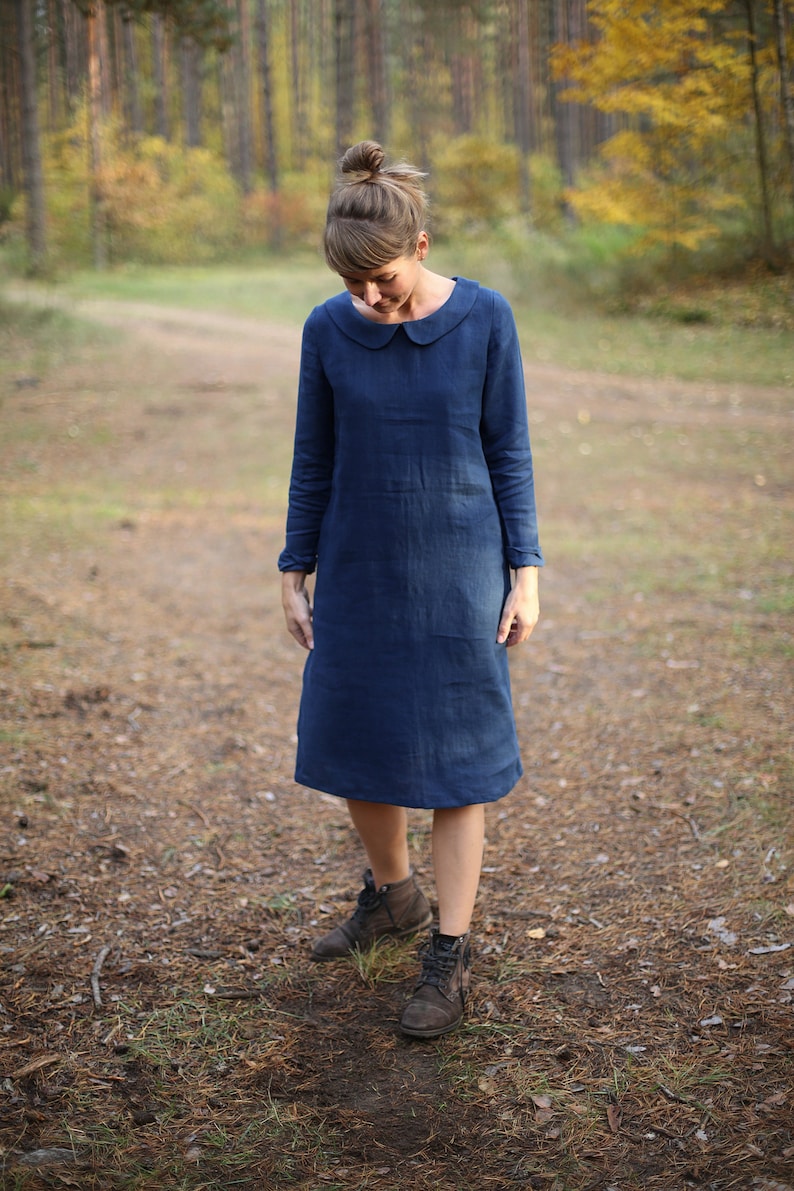May include: A long-sleeved, navy blue dress with a Peter Pan collar. The dress falls to the mid-calf and is worn with brown ankle boots. The person is standing on a forest path.