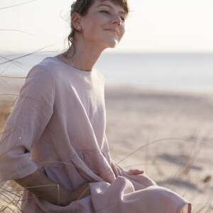 May include: A woman wearing a light pink linen dress sits in a field of tall grass. She is looking up and smiling.