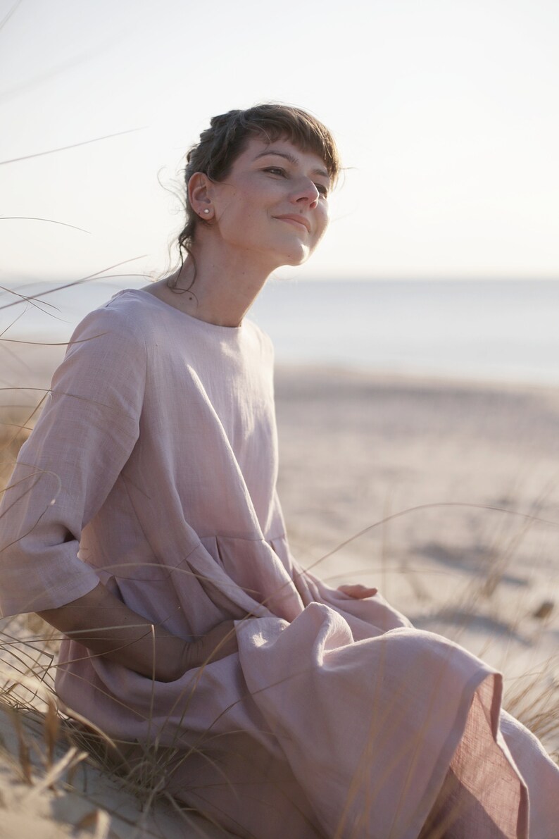 May include: A woman wearing a light pink linen dress sits in a field of tall grass. She is looking up and smiling.
