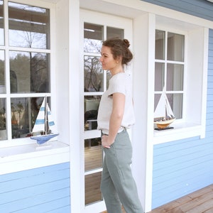 May include: A woman wearing a light beige top and olive green pants stands in front of a blue and white house. She is wearing brown boat shoes and has her hands in her pockets.