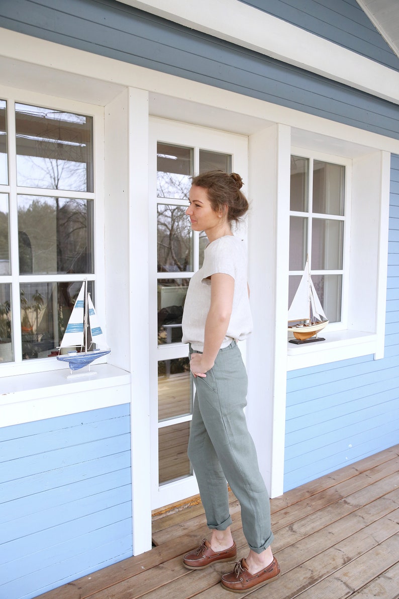 May include: A woman wearing a light beige top and olive green pants stands in front of a blue and white house. She is wearing brown boat shoes and has her hands in her pockets.
