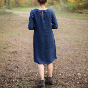 May include: A long-sleeved, navy blue dress with a Peter Pan collar and a keyhole back detail. The dress falls to mid-calf and is worn with brown ankle boots. The background features a forest with a path.
