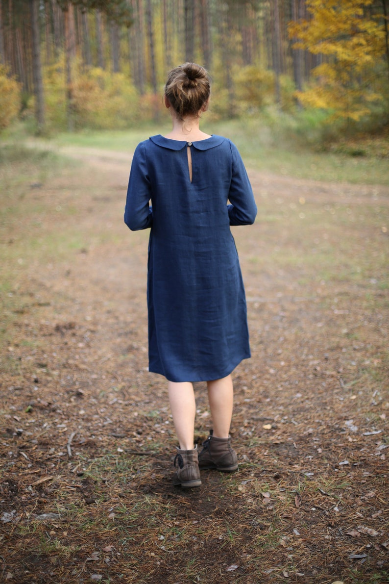 May include: A long-sleeved, navy blue dress with a Peter Pan collar and a keyhole back detail. The dress falls to mid-calf and is worn with brown ankle boots. The background features a forest with a path.