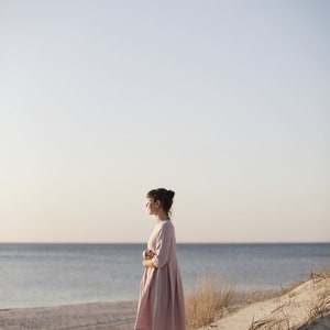 May include: A woman in a light pink dress stands on a sandy beach with the ocean in the background.