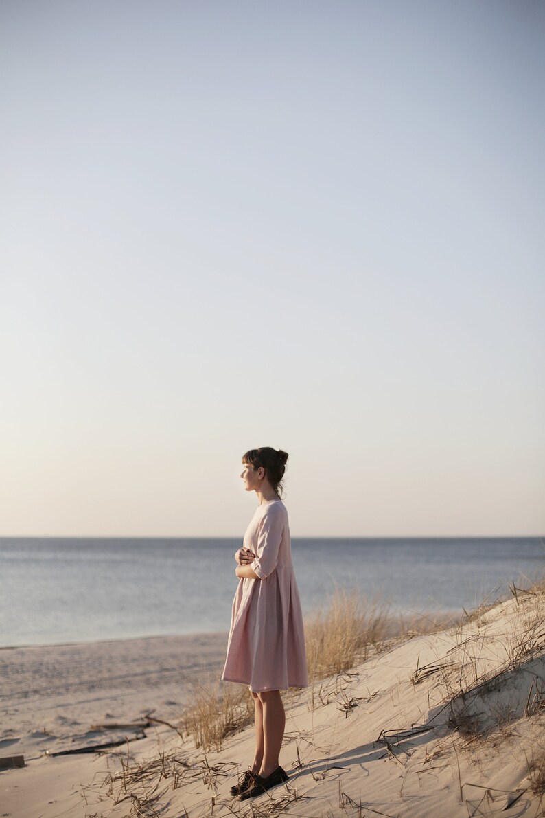 May include: A woman in a light pink dress stands on a sandy beach with the ocean in the background.