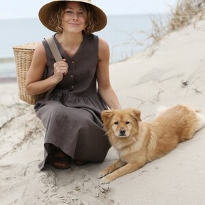 May include: A woman wearing a straw hat and a brown linen dress sits on a sandy beach with a wicker basket and a golden brown dog.