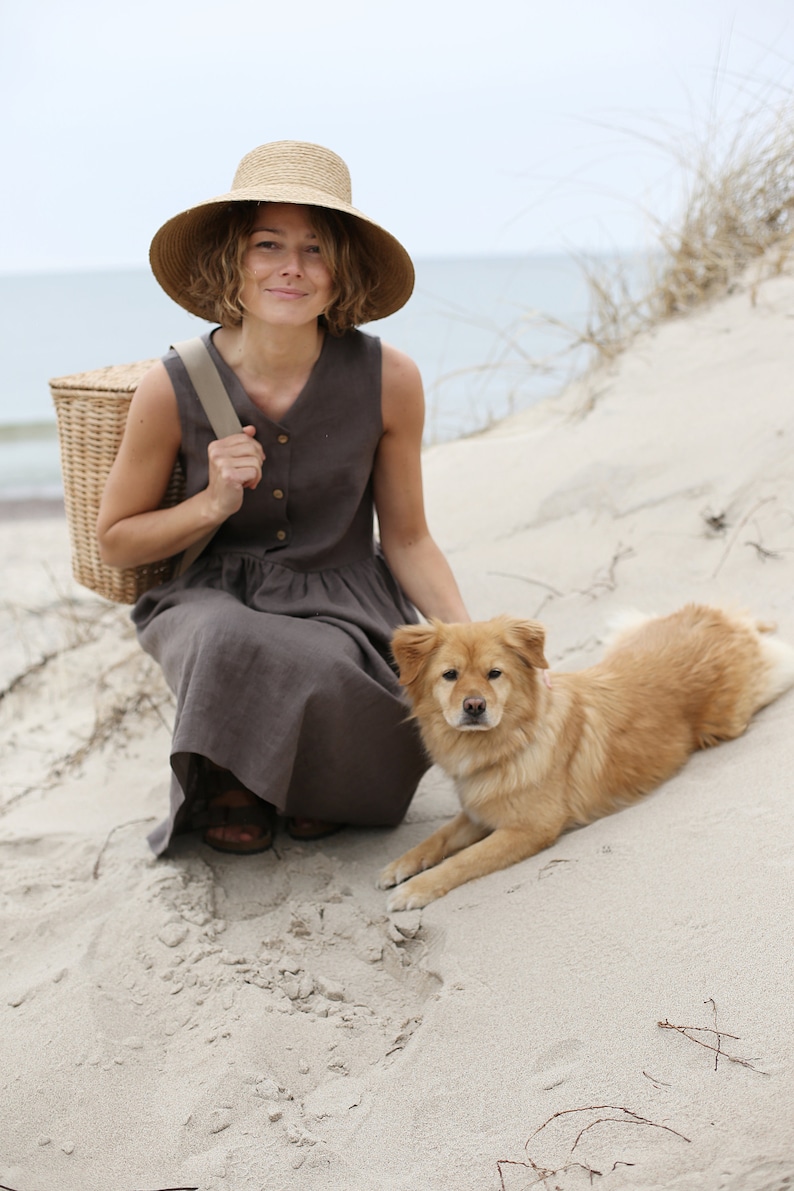 May include: A woman wearing a straw hat and a brown linen dress sits on a sandy beach with a wicker basket and a golden brown dog.