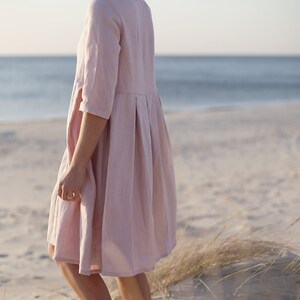 May include: A woman wearing a light pink linen dress stands on a sandy beach with tall grass in the foreground. The dress has a pleated skirt and 3/4 sleeves.
