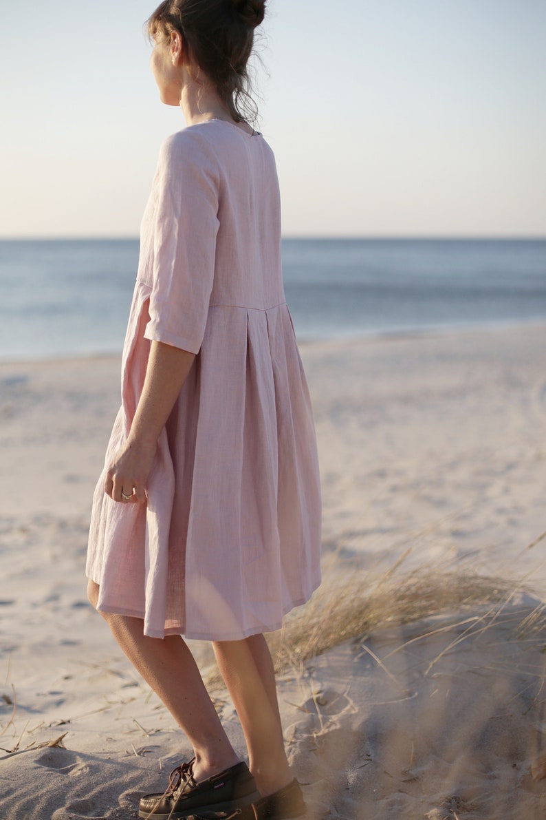May include: A woman wearing a light pink linen dress stands on a sandy beach with tall grass in the foreground. The dress has a pleated skirt and 3/4 sleeves.