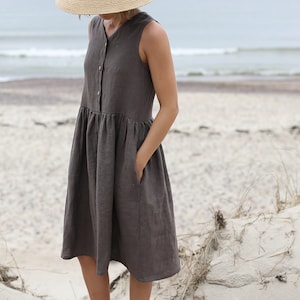 May include: A woman wearing a brown linen dress with a wide-brimmed straw hat and brown sandals. She is standing on a sandy beach.