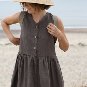 May include: A woman wearing a brown linen dress with a button-up front and a wide brimmed straw hat. The dress has a gathered skirt and is sleeveless. The woman is standing on a sandy beach.