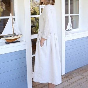 Pu&ograve; includere: Una donna indossa un abito di lino beige chiaro con una gonna ampia a pieghe e un cappello di paglia con un nastro nero. &Egrave; in piedi su un ponte di legno di fronte a una casa bianca con rivestimento blu.