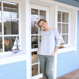 May include: A woman wearing a light gray top and green pants stands in front of a light blue house with white trim. She is smiling and has her hands in her pockets. She is wearing brown boat shoes.