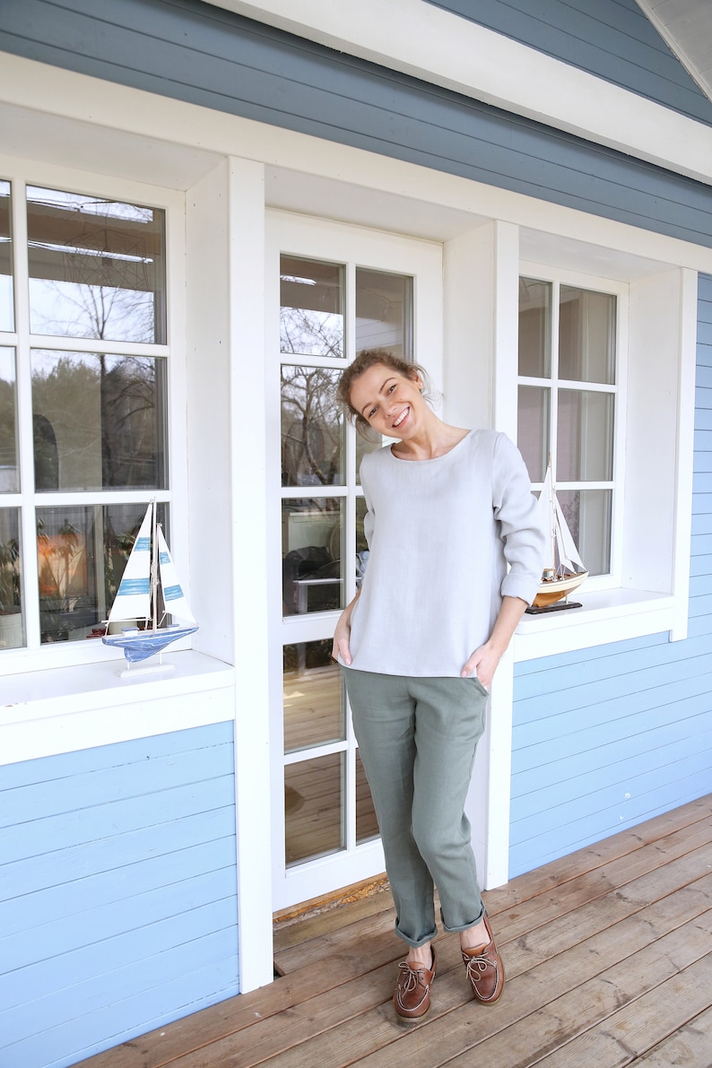 May include: A woman wearing a light gray top and green pants stands in front of a light blue house with white trim. She is smiling and has her hands in her pockets. She is wearing brown boat shoes.