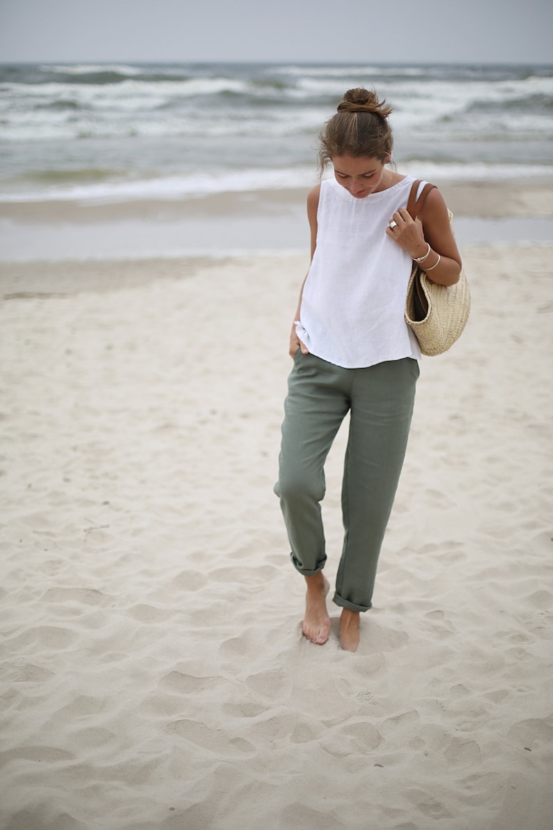 May include: A woman wearing a white sleeveless top and olive green pants walks on a sandy beach. She is carrying a straw tote bag with a brown leather handle.