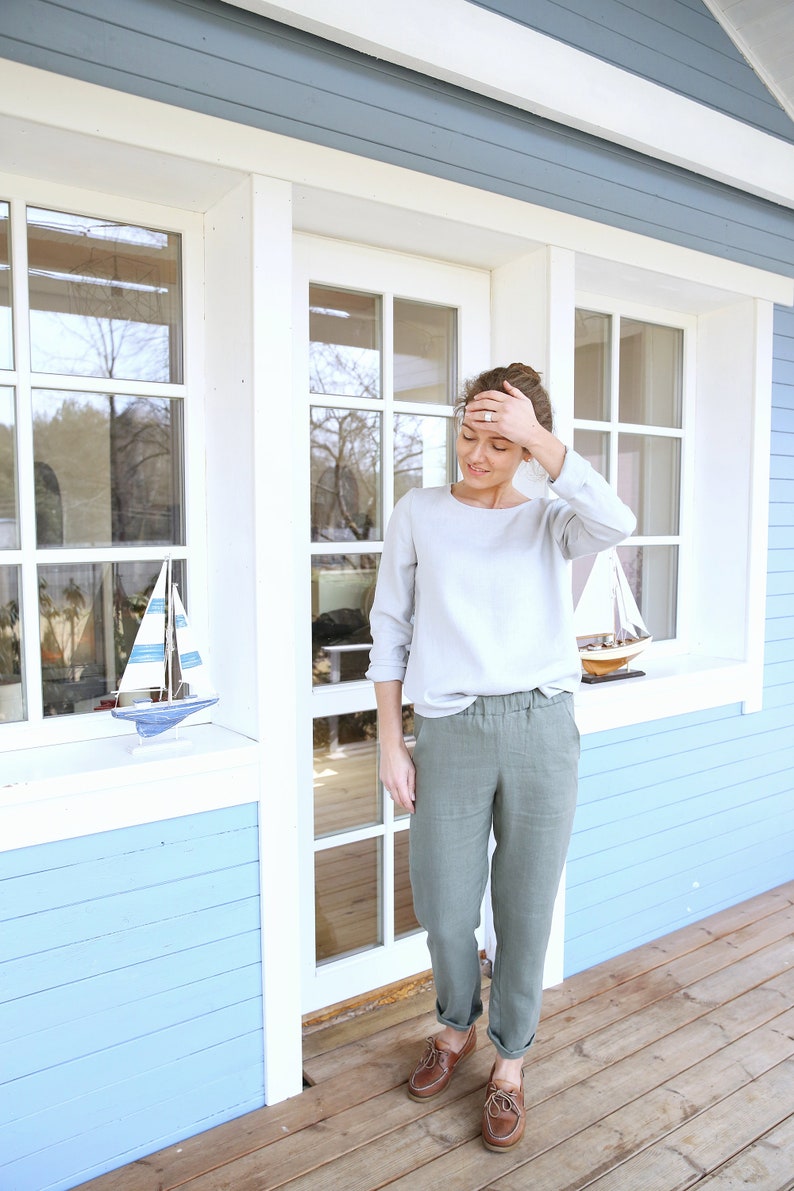 May include: A woman wearing a light gray long-sleeved shirt and olive green pants stands in front of a blue house with white trim. She is wearing brown boat shoes and has her hand on her forehead.