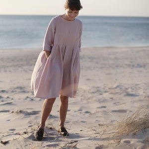 May include: A woman wearing a light pink linen dress walks on a sandy beach. The dress has a loose fit and a gathered skirt.