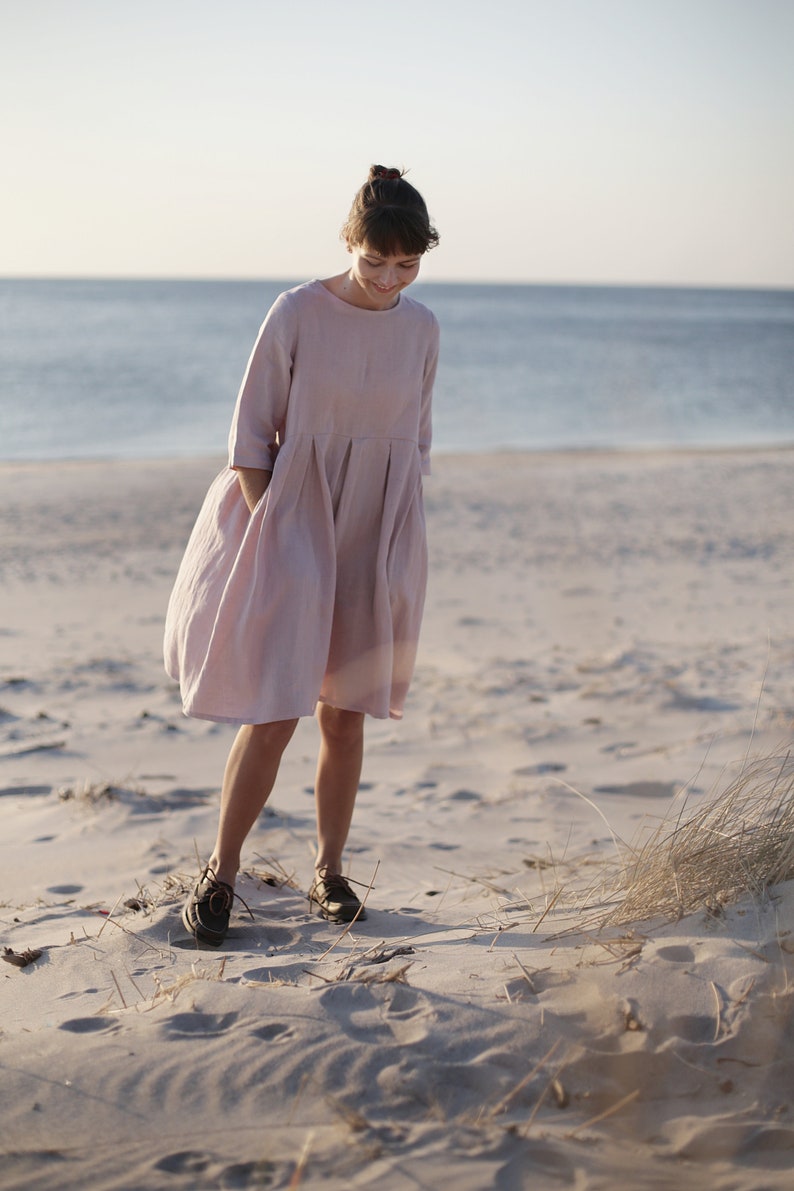 May include: A woman wearing a light pink linen dress walks on a sandy beach. The dress has a loose fit and a gathered skirt.